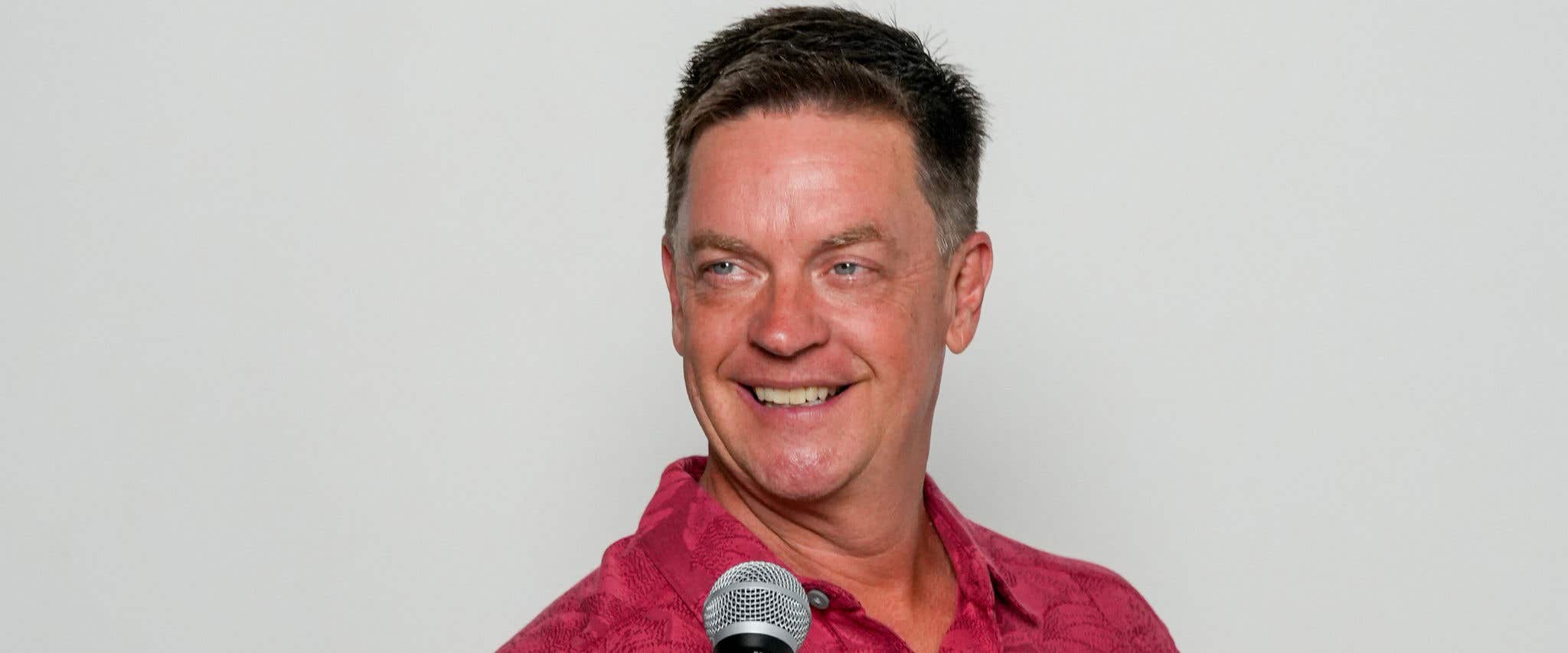 Jim Breuer in red Hawaiian shirt holding a microphone looking and smiling into the distance. 