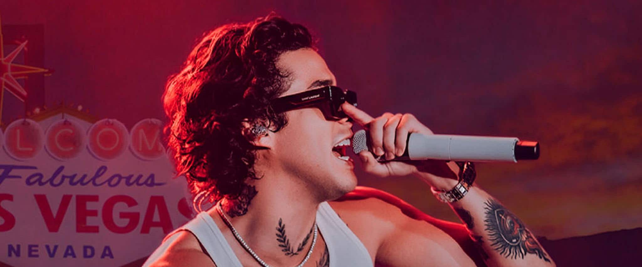 Mario Bautista in a white tank and black bottoms holding a microphone and pushing up dark sunglasses standing in front of Welcome to Fabulous Las Vegas sign.     