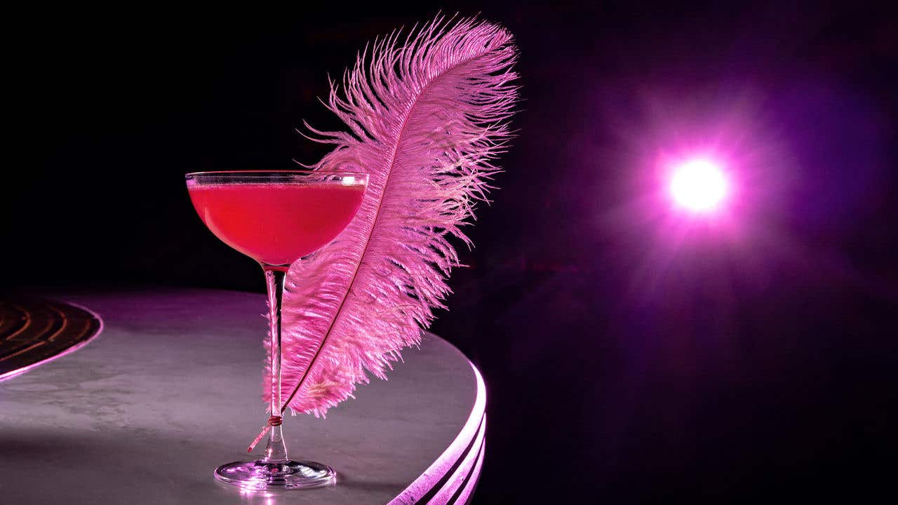 Pink cocktail in a coupe glass with a large pink feather garnish, lit by a bright pink spotlight.