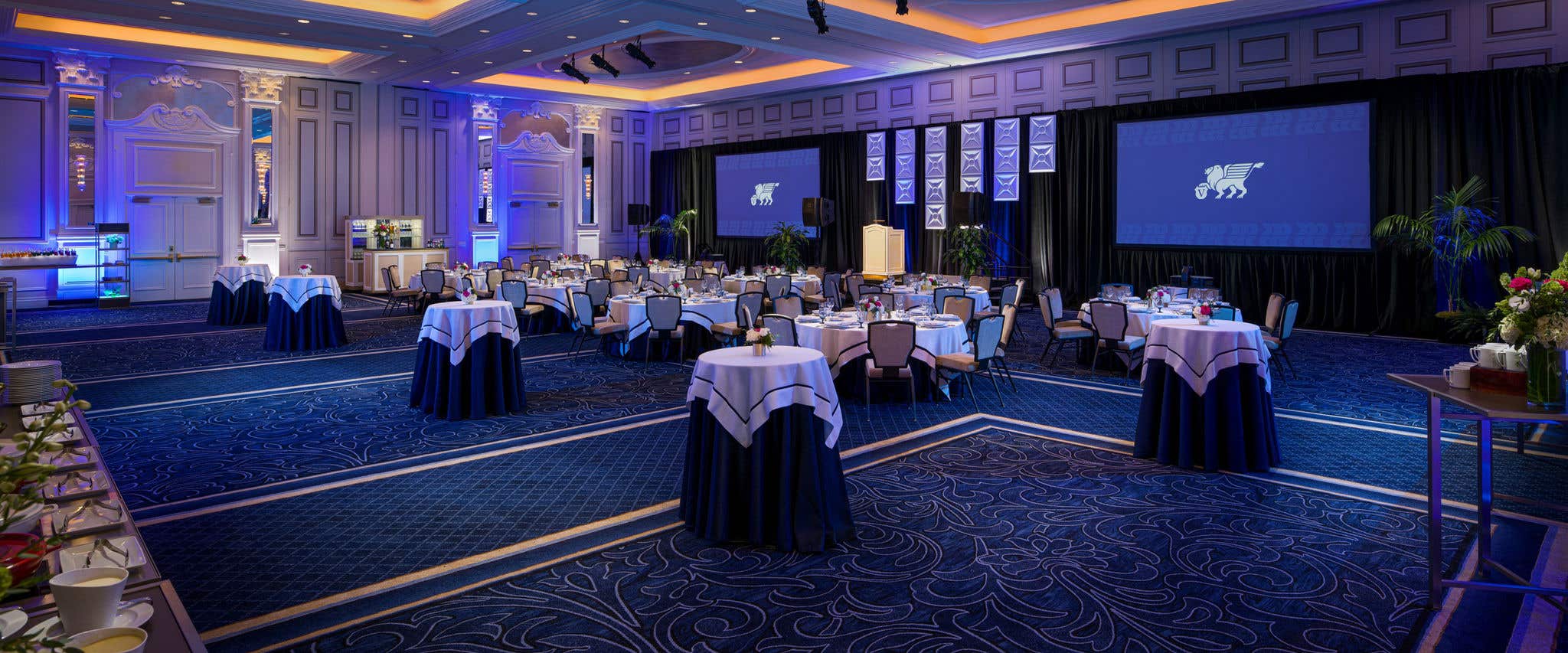 Elegant ballroom with round tables in navy and white linens, blue accent lighting, two projection screens, and a staged podium.