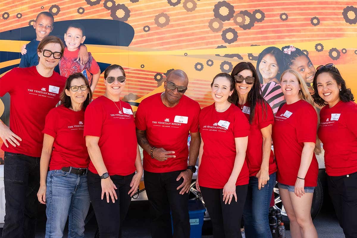 A group of eight smiling people wearing matching red shirts pose for a group photo in front of a colorful mural.