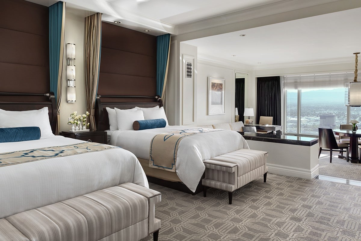 Luxurious hotel room with two double beds, a seating area, and a large window offering a city view.