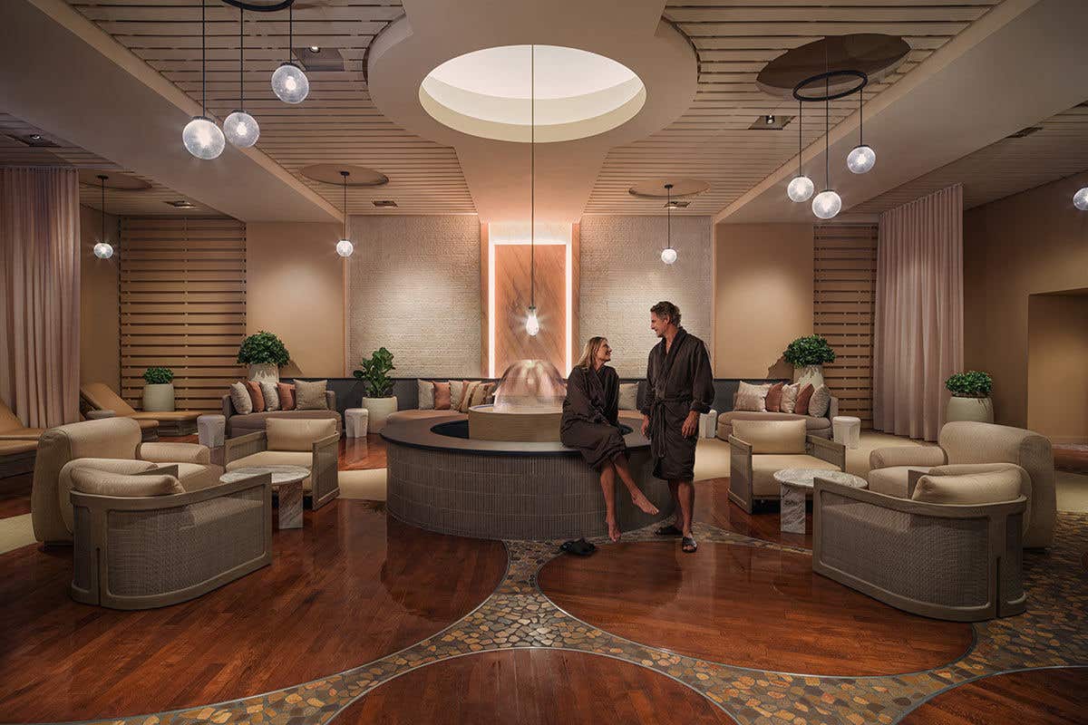 A man and a woman in dark grey spa robes relax in a spacious, circular lounge area with modern decor. They are sitting on the edge of a central water feature, engaged in conversation. The room features warm wood floors with stone mosaic details, comfortable armchairs, and plush sofas against a slatted wood wall. Soft, ambient light comes from spherical pendant lights and recessed ceiling fixtures, creating a tranquil and luxurious atmosphere.