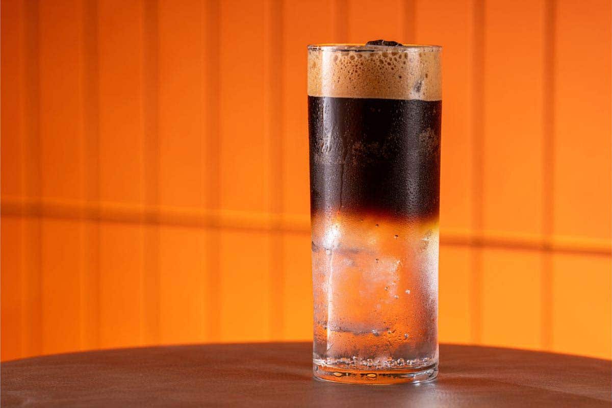 A cocktail in a tall glass, with brown liquor on top and clear liquor on the bottom on a table with an orange background.