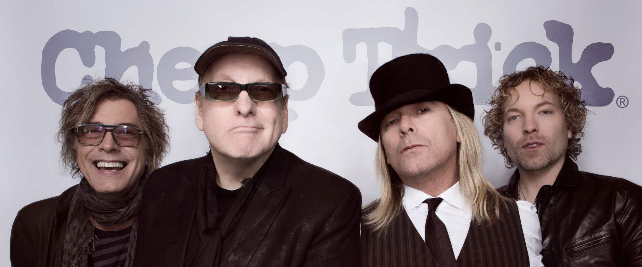 Cheap Trick the band standing in front of a wall with the band’s name in the background looking at the camera. 