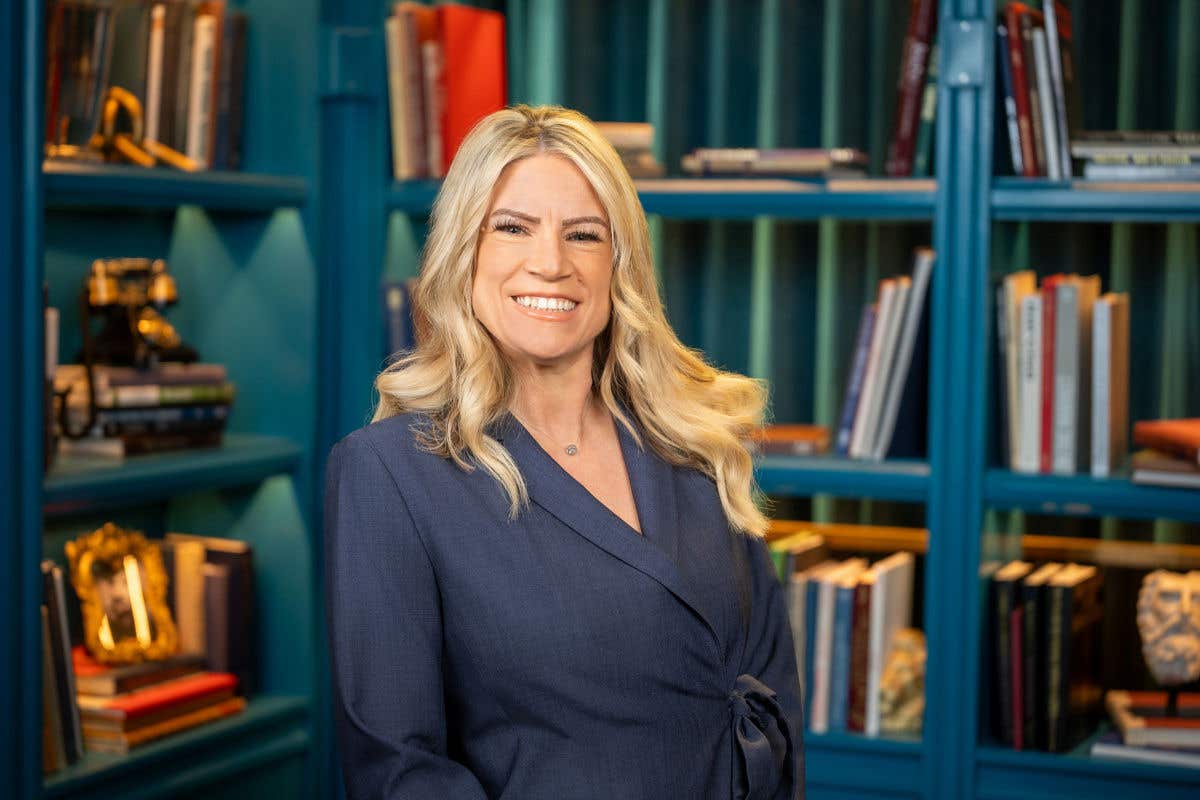 A professional headshot of a smiling woman with long blonde hair, wearing a dark blue wrap-style blazer. She is posed in front of a teal bookshelf background featuring colorful books and vintage decor.