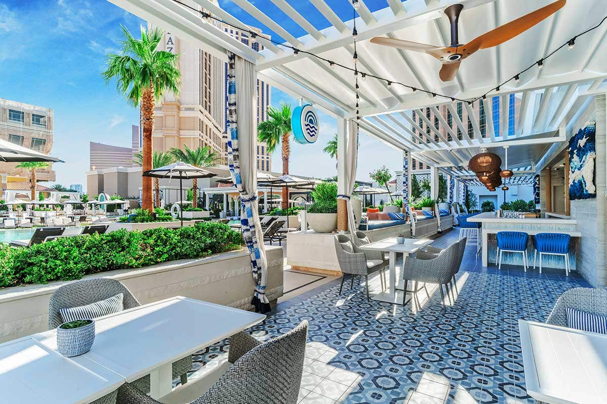 A covered open air restaurant dining room on a pool deck with tables, chairs, and a bar.
