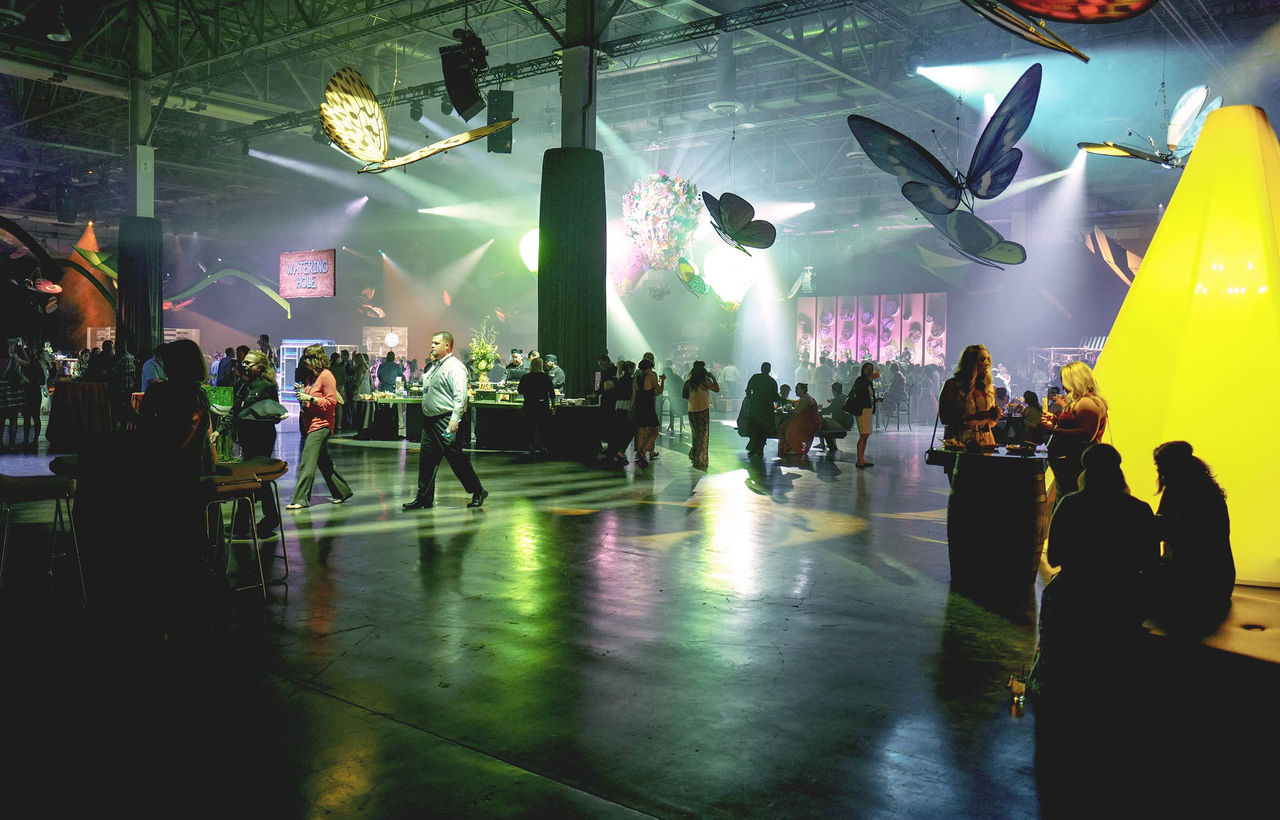 Dark lit expo hall with large butterfly cutouts hanging from the ceiling and people mingling. 